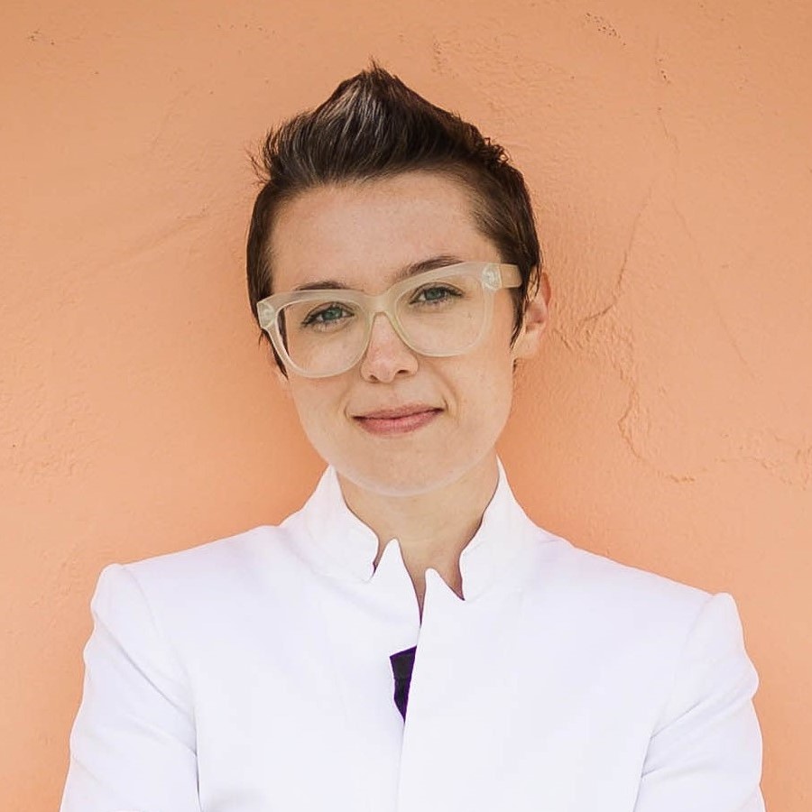 MPH student Madison Saunders stands against a peach-colored wall. Her short hair is spiked and she's wearing large glasses