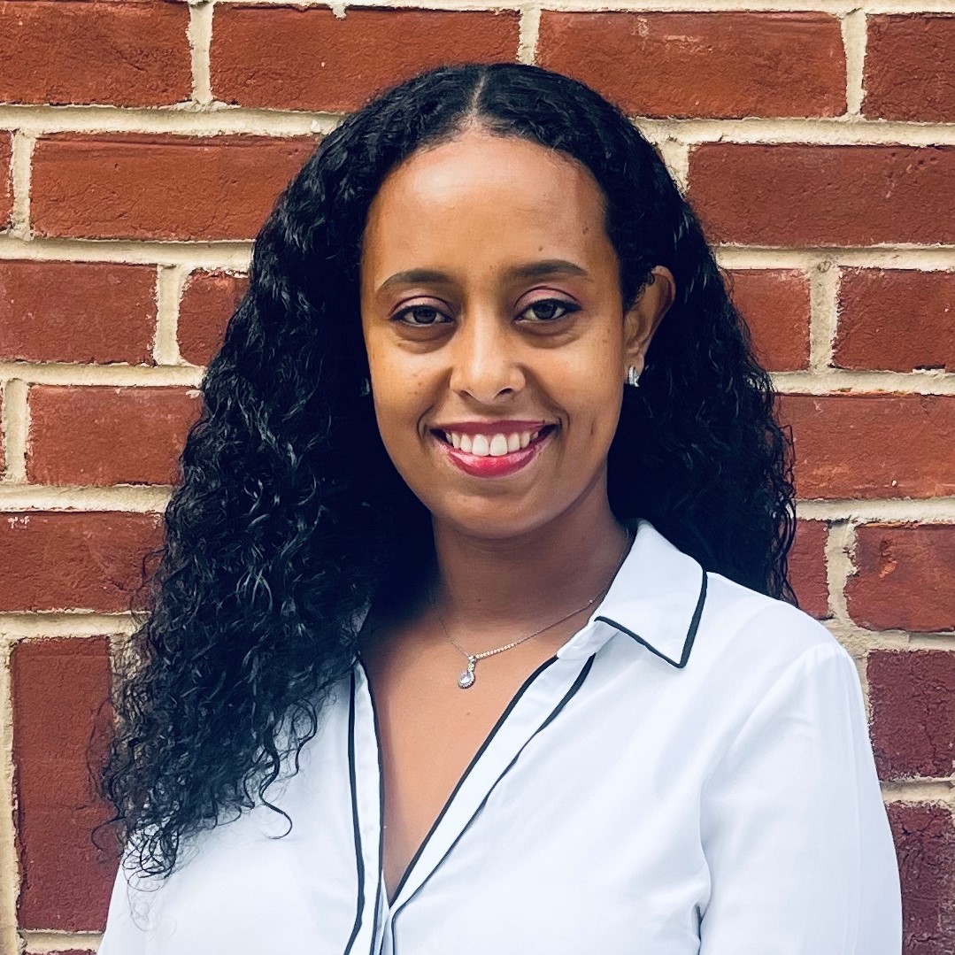 Dr Tsion Gebre stands in front of a red brick wall. She is wearing a white lab coat and her dark hair is down. 