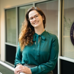 Amelia Wallace, PhD, leans on a railing in an atrium. She is wearing a green shirt and has long brown hair swept over one shoulder. 