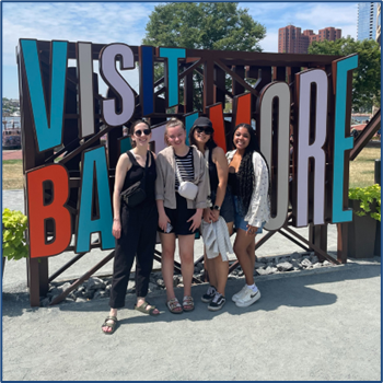 Four 2024 CHAMP trainees stand in front of a colorful sign that reads Visit Baltimore