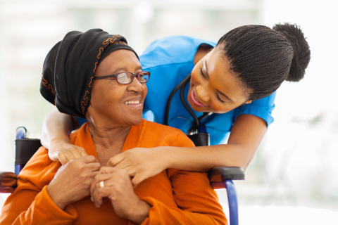 A healthcare worker leans over the shoulders of a seated older person. They are smiling at each other.