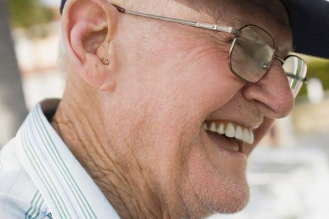 An older, bespectacled white man smiles in profile, showing his hearing aid